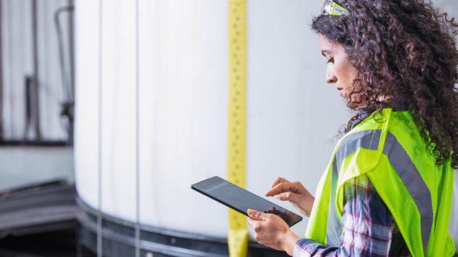 Female worker wearing neon vest and safety glasses using tablet. Industrial vats visible in background.