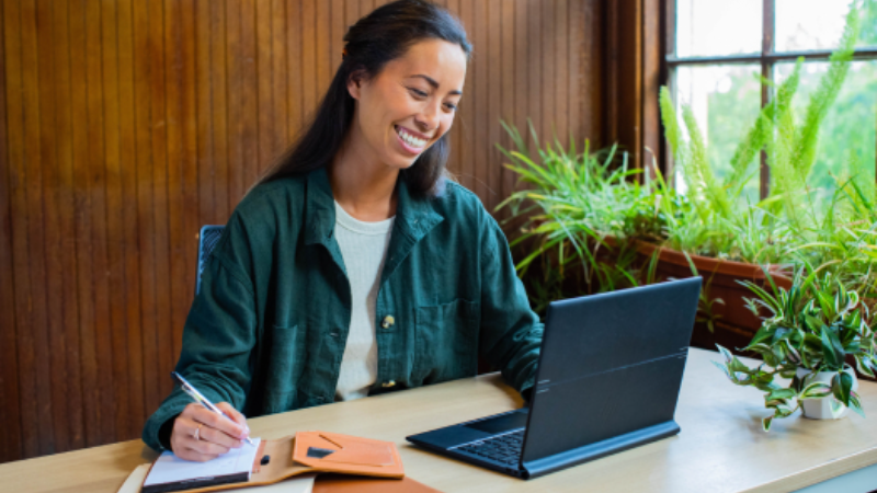 Woman working on a laptop and taking notes in a bright and natural workspace.