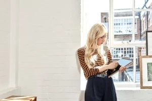 A woman sits in a comfortable chair in her bright sun-filled apartment using her Lenovo Yoga computer in tablet mode.