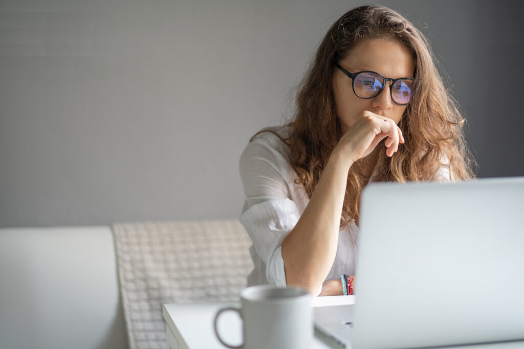 Young n a white shirt and glasses sits at a table in front of a laptop, looks seriously at the screen, thinks intently