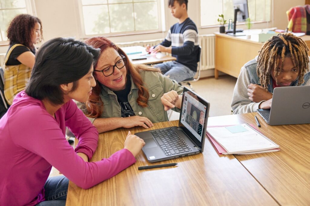 A classroom scene with students at communal desks and an educator leans in to help one of the students.