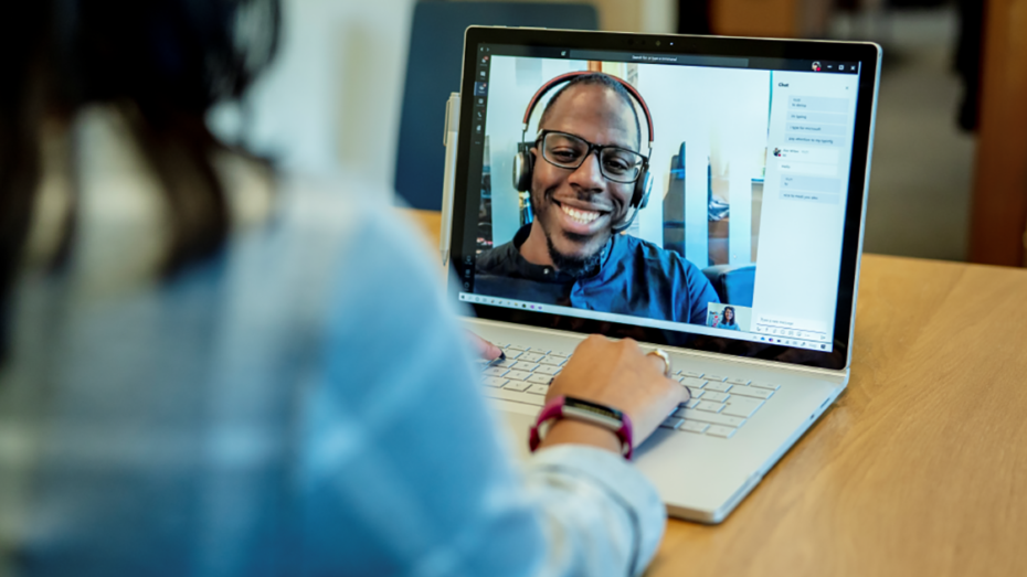 Image of a woman at a desk using a Surface laptop to make a Microsoft Teams video call with one man smiling and wearing a headset.