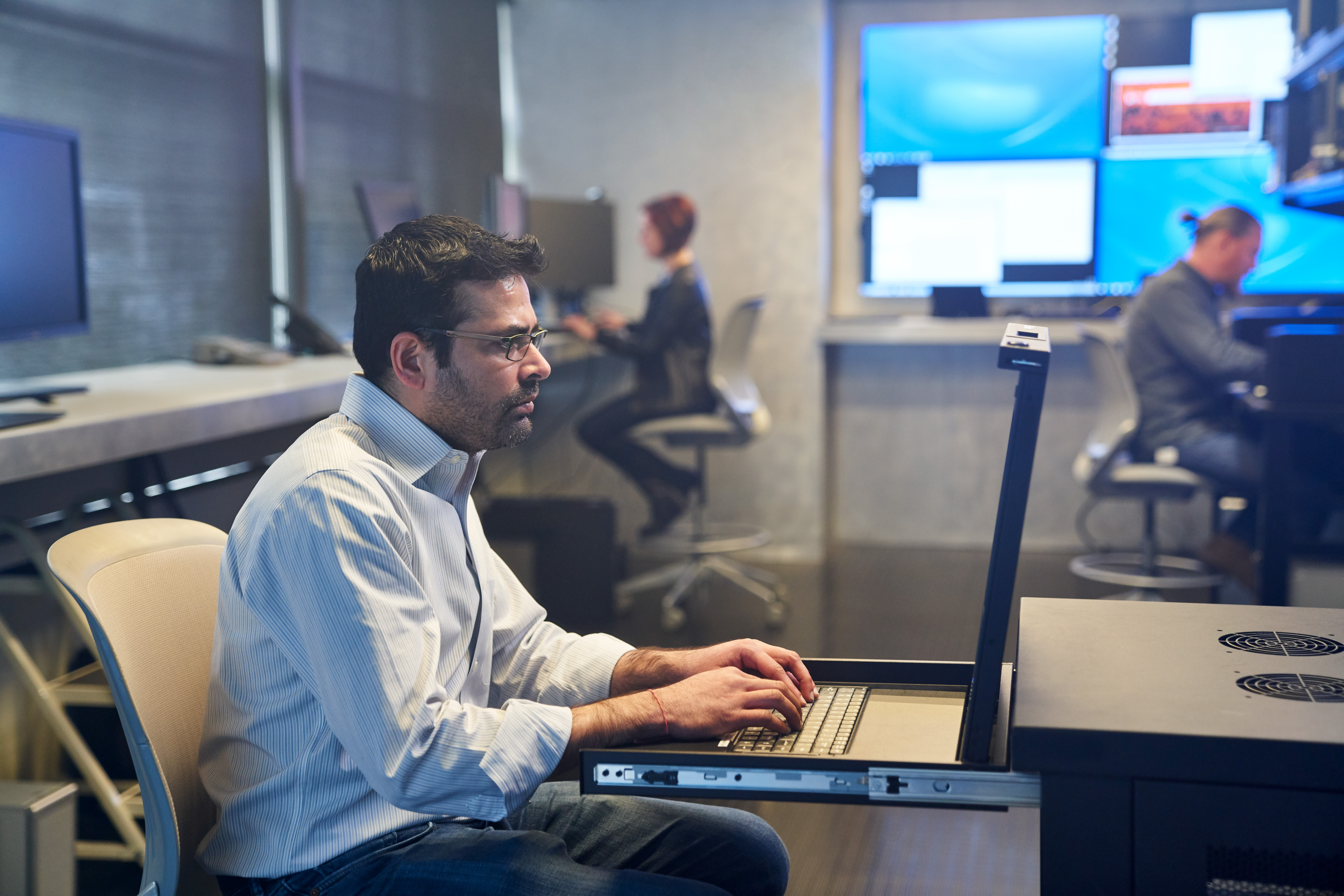 Man in a collared shirt working on a server station inside a secure room. Coworkers and large monitors are in the background. Keywords: network security, on-premise security, threat protection, secure score, monitoring, security management, operations, Microsoft Security collection