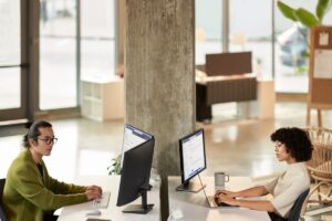 A man and a woman collaborating during a Microsoft Teams meeting while working in an open office setting on dual monitor​s.