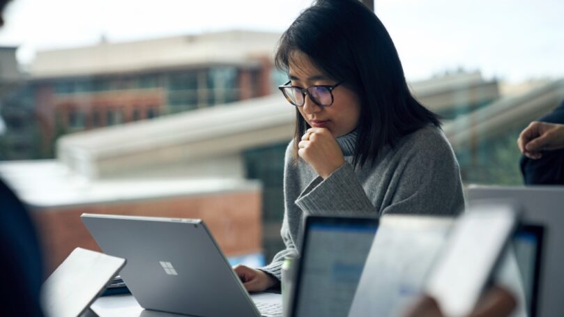 Woman with glasses sitting at a community table in common area reading on a Surface laptop