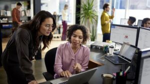 Two employees collaborate over a computer in an open office workspace.